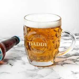 Puede incluir: Un vaso de cerveza transparente lleno de cerveza y espuma, con las palabras "DADDY OUR HERO" en un diseño circular. El nombre "JOHN DAVIES" está encima de la palabra "DADDY". Una botella de cerveza marrón está en el fondo.