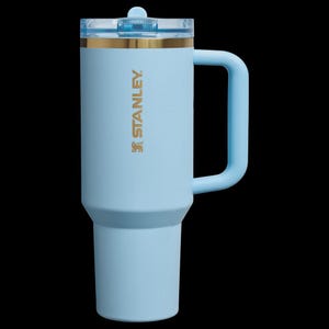 May include: A light blue Stanley insulated tumbler with a handle and a clear lid. The tumbler has a gold band near the top and the Stanley logo in gold. The handle and lid are also light blue.