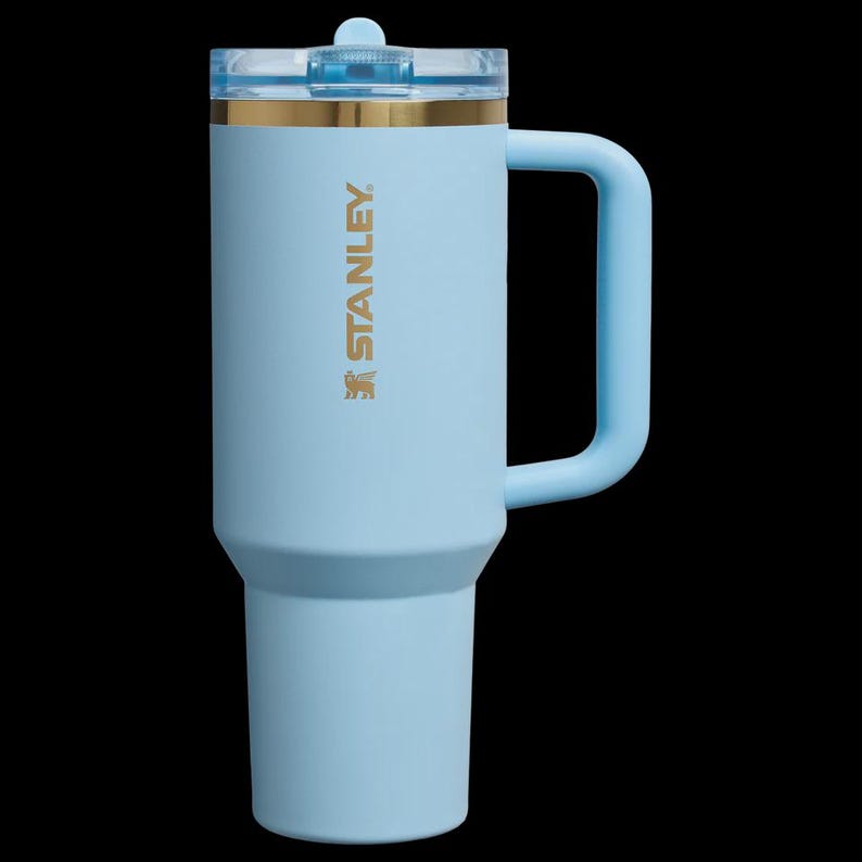 May include: A light blue Stanley insulated tumbler with a handle and a clear lid. The tumbler has a gold band near the top and the Stanley logo in gold. The handle and lid are also light blue.