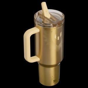May include: A gold-colored insulated tumbler with a cream-colored handle and lid. The tumbler features a heart-shaped design with sunburst rays and a logo. The lid has a built-in straw. The tumbler is designed to keep beverages hot or cold.
