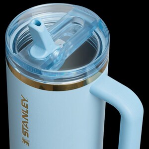 May include: A light blue Stanley mug with a clear lid and a matching blue handle. The mug has a gold band near the top and the Stanley logo is visible on the side. The lid features a flip-top opening for easy access.