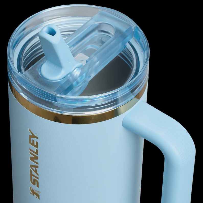 May include: A light blue Stanley mug with a clear lid and a matching blue handle. The mug has a gold band near the top and the Stanley logo is visible on the side. The lid features a flip-top opening for easy access.