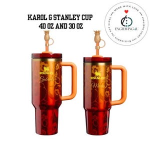 Op de afbeelding: Twee Stanley-bekers, 1,18L en 887ml, met oranje handvatten en ananas-strohalmen. De bekers zijn rood en goud met bloemmotieven en de namen "Gabriela" en "Maria". De tekst "KAROL G STANLEY CUP" staat bovenaan.
