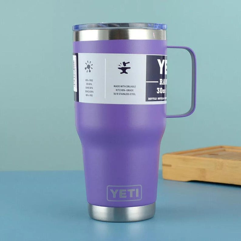May include: A purple 30 oz Yeti Rambler mug with a handle. The mug has a stainless steel base and rim, a clear lid, and a white label with text and icons. The Yeti logo is displayed on the front.