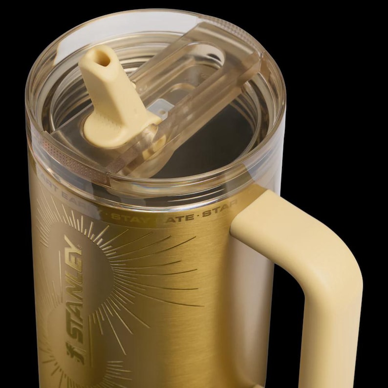 May include: A gold-colored Stanley travel mug with a cream-colored handle and lid. The clear lid has a cream-colored spout. The mug has the Stanley logo and the words "Stay" and "Star" etched on the side.