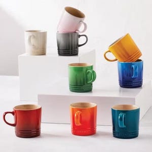 以下が含まれることがあります： 赤、オレンジ、緑、青、黄色、ピンク、グレーなど、さまざまな色合いのカラフルなセラミックマグのコレクション。各マグには対照的な内装と湾曲したハンドルが付いています。マグは白いプラットフォームに配置されています。