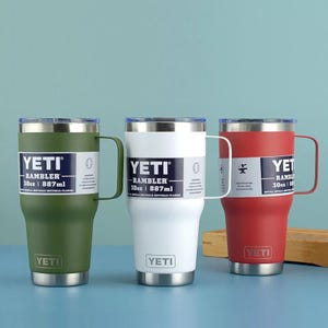 May include: Three Yeti Rambler 30oz (887ml) insulated mugs in green, white, and red. Each mug has a silver handle and a label with the Yeti logo and product information. The mugs are designed for beverages.