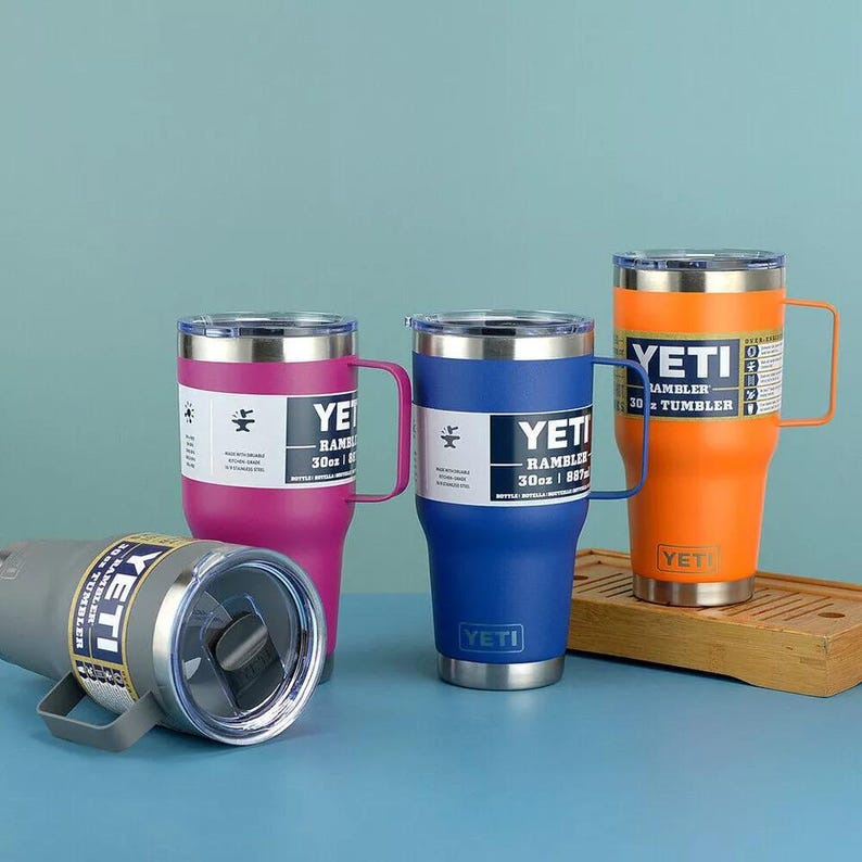 May include: A collection of Yeti Rambler 30 oz tumblers in various colors: gray, pink, blue, and orange. Each tumbler has a handle, a clear lid, and a stainless steel base. The Yeti logo is visible on each tumbler.