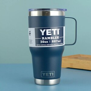 May include: A navy blue YETI Rambler mug with a handle, stainless steel accents, and a clear lid. The mug has a capacity of 30oz (887ml) and features the YETI logo.