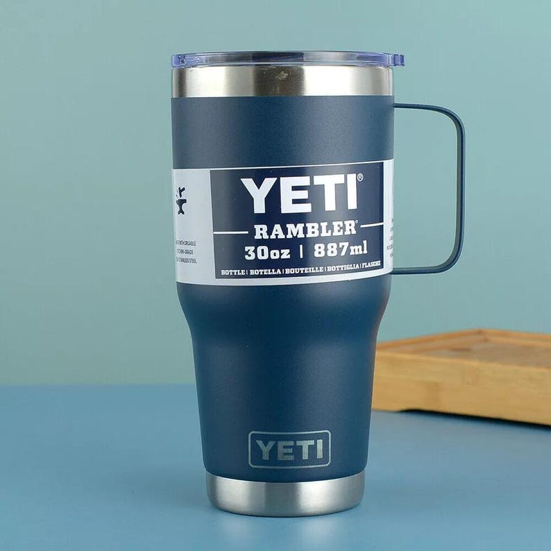 May include: A navy blue YETI Rambler mug with a handle, stainless steel accents, and a clear lid. The mug has a capacity of 30oz (887ml) and features the YETI logo.