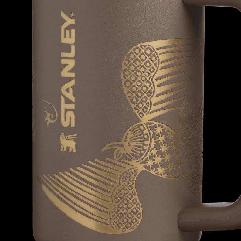 Puede incluir: Taza Stanley marr&oacute;n con dise&ntilde;o de p&aacute;jaro dorado. La taza tiene un asa y el logotipo de Stanley est&aacute; impreso verticalmente en el lateral. El dise&ntilde;o del p&aacute;jaro est&aacute; detallado con patrones geom&eacute;tricos.