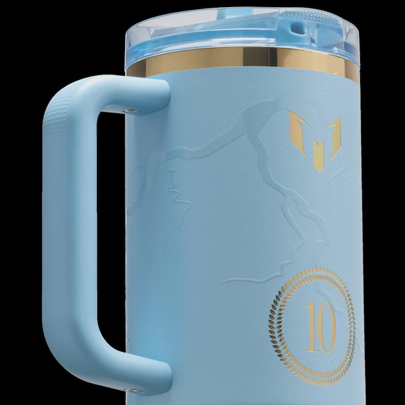 May include: A light blue insulated mug with a handle and a clear lid. The mug features gold accents and a gold emblem. The number "10" is displayed in gold within a circular design. The mug is designed to hold approximately 24 ounces.