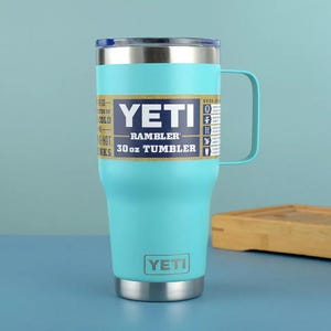 May include: A teal YETI Rambler 30 oz tumbler with a silver handle and rim. The tumbler has a white and blue label with the YETI logo and product information. The tumbler is designed to keep drinks cold or hot.