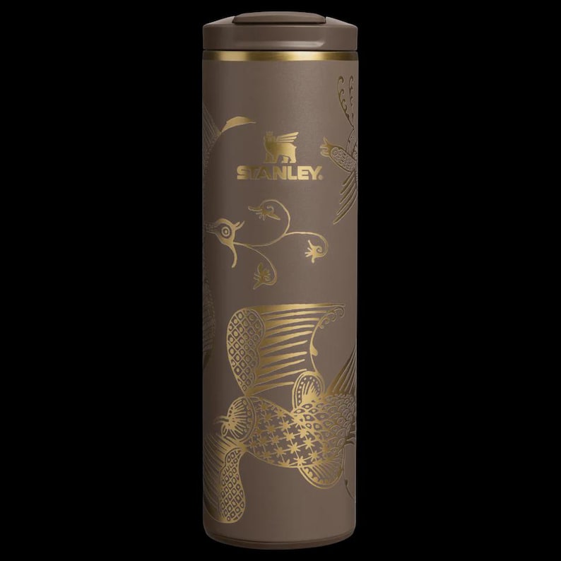 Puede incluir: Un termo de viaje Stanley marr&oacute;n con dise&ntilde;os dorados de p&aacute;jaros y flores. El termo tiene una tapa marr&oacute;n y una banda dorada en la parte superior. El logotipo de Stanley es visible en la parte delantera.