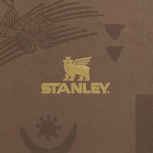 Puede incluir: Un fondo marr&oacute;n con un emblema dorado que presenta un oso con alas y una corona, con la palabra "STANLEY" debajo. La imagen tambi&eacute;n incluye un p&aacute;jaro y otros elementos decorativos.