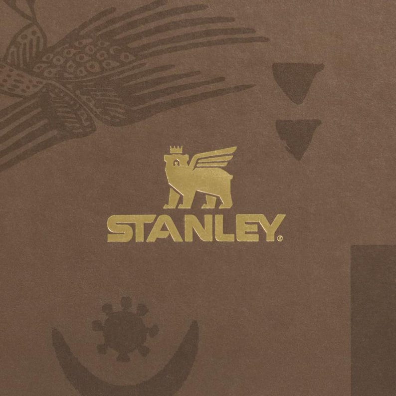 Puede incluir: Un fondo marr&oacute;n con un emblema dorado que presenta un oso con alas y una corona, con la palabra "STANLEY" debajo. La imagen tambi&eacute;n incluye un p&aacute;jaro y otros elementos decorativos.