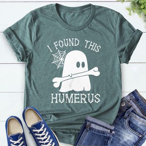 May include: A heather forest green t-shirt with a white graphic of a ghost holding a bone, with the text "I FOUND THIS HUMERUS". Blue denim jeans and blue sneakers are also in the image.