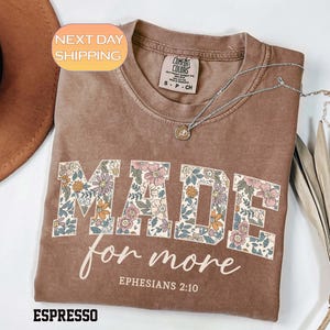 May include: Espresso-colored t-shirt with the words "MADE for more" in a floral design. The text "Ephesians 2:10" is below the main text. A silver necklace with a pendant rests on the shirt. The shirt is folded and has a Comfort Colors tag.