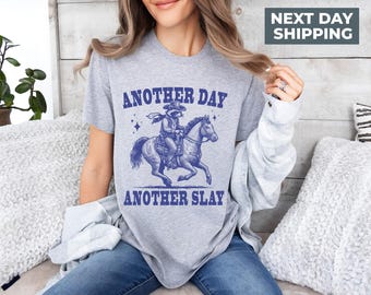 May include: A gray t-shirt with a blue graphic of a raccoon riding a horse. The graphic says "Another Day Another Slay".