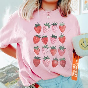 May include: A light pink crewneck t-shirt featuring a repeating grid pattern of sixteen strawberries in various shades of red and pink. The shirt has short sleeves and a round neckline. A vertical orange tag on the right side reads COMFORT COLORS.