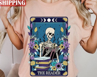 May include: A light peach-colored t-shirt featuring a tarot card design with a skeleton reading a book. The card is framed with purple, blue, and yellow flowers. The text "THE READER" is at the bottom of the card.