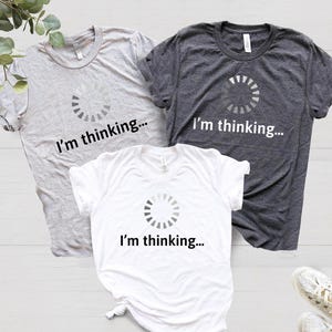 May include: Three t-shirts with a loading circle graphic and the text "I'm thinking..." printed on them. The shirts are in grey, white, and dark grey.