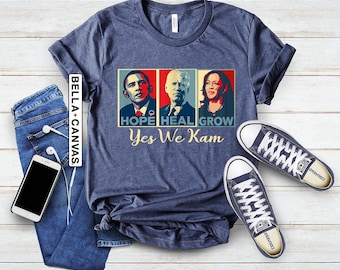 Pode incluir: Uma camiseta azul marinho com um gráfico de três rostos de pessoas. O texto na camiseta diz "Hope Heal Grow Yes We Kam".
