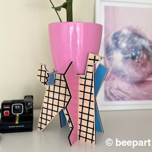 May include: A pink plant pot sits on a geometric wooden stand with black, pink, and blue accents. The stand is made of plywood and has a grid pattern.
