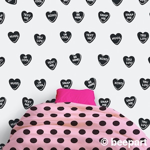 May include: Black and white heart-shaped stickers with various messages such as "Text Me", "Be Mine", "Hugs", "Snap Me", "Tru Luv", "Ur Cool", and "XOXO". The stickers are on a white wall behind a pink bed with black polka dots.