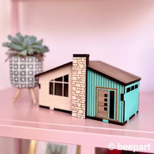 May include: A miniature wooden model of a mid-century modern house with a teal blue exterior, brown roof, and a stone chimney. The house has a single window and a door with a wood grain finish.
