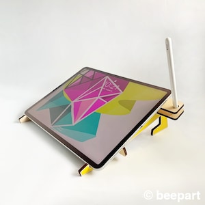 May include: A yellow wooden stand with a tablet propped up on it. The tablet has a colorful abstract design on the screen. A white stylus is placed in a slot on the stand.