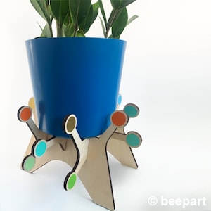 Puede incluir: Una maceta de cerámica azul con una planta que crece en ella se asienta sobre un soporte de madera con círculos coloridos. El soporte tiene un diseño único con múltiples patas.