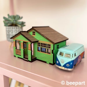 May include: A green wooden model house with a brown roof and a blue and white toy van parked in front of it. The house has a porch and windows. The model is sitting on a pink surface.