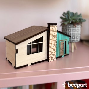 Puede incluir: Un modelo de casa moderno de mediados de siglo en miniatura de madera con techo marrón, paredes blancas, una puerta azul turquesa y una chimenea de piedra.