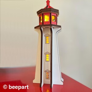 May include: A wooden lighthouse with a red top and brown trim. The lighthouse has three windows with a yellow glow, and the top section has multiple windows. The base is white, and the lighthouse is on a red surface.