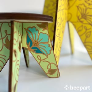 May include: A small, green and gold painted wooden stool with a floral design. The stool has a round top and three legs.