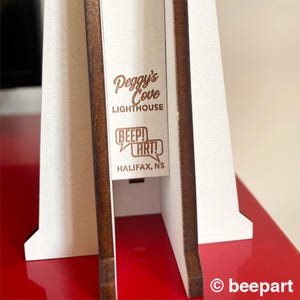 May include: A wooden lighthouse model with white and brown accents. The text "Peggy's Cove Lighthouse" and "BEEP! ART!" are engraved on the front. The base is red, and the design includes the text "HALIFAX, NS".