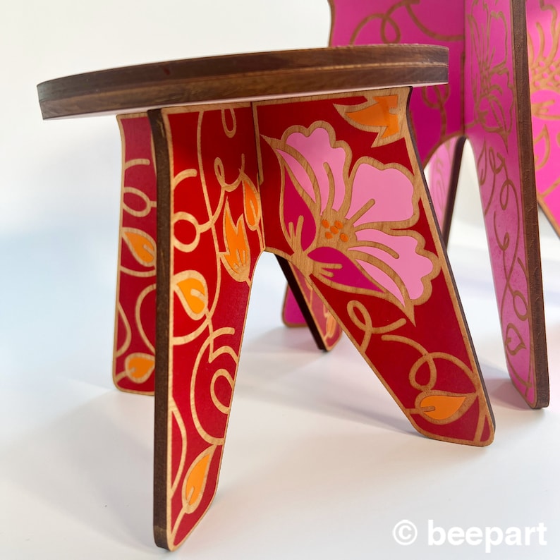 May include: A small wooden stool with a red and pink floral design. The stool has a round top and three legs.