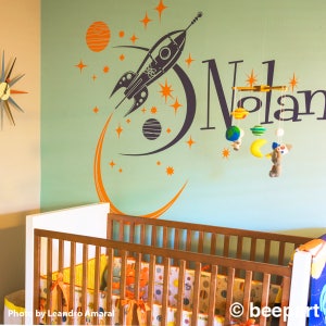 May include: A nursery room with a crib and a wall decal featuring a rocket ship, planets, and stars. The name "Nolan" is written in black lettering on the wall.