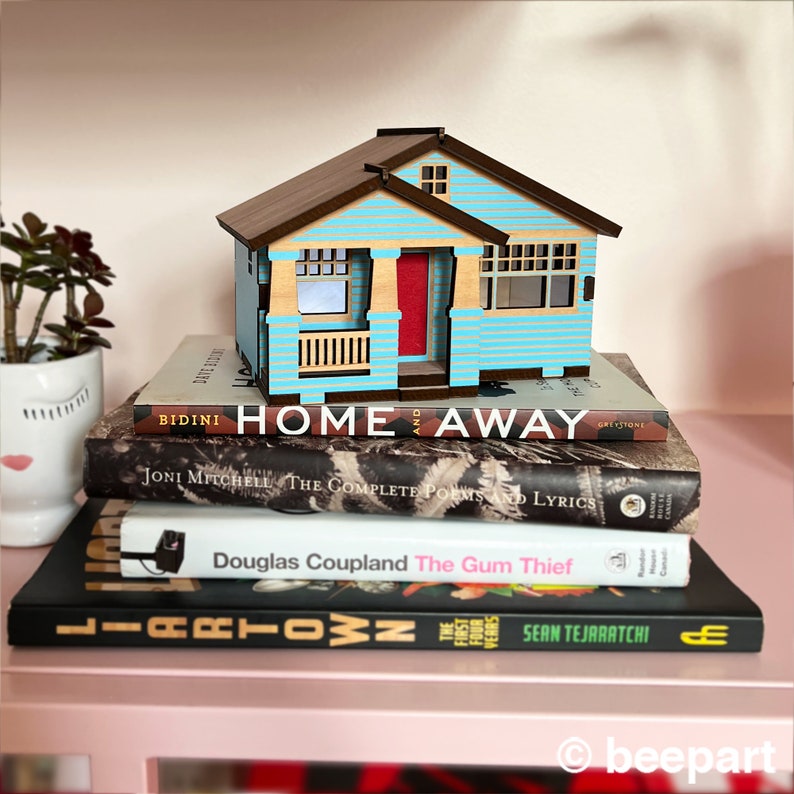 May include: A wooden model of a light blue house with a red door, sitting on top of a stack of books. The books are titled "Home Away", "The Complete Poems and Lyrics", "The Gum Thief", and "Liartown".