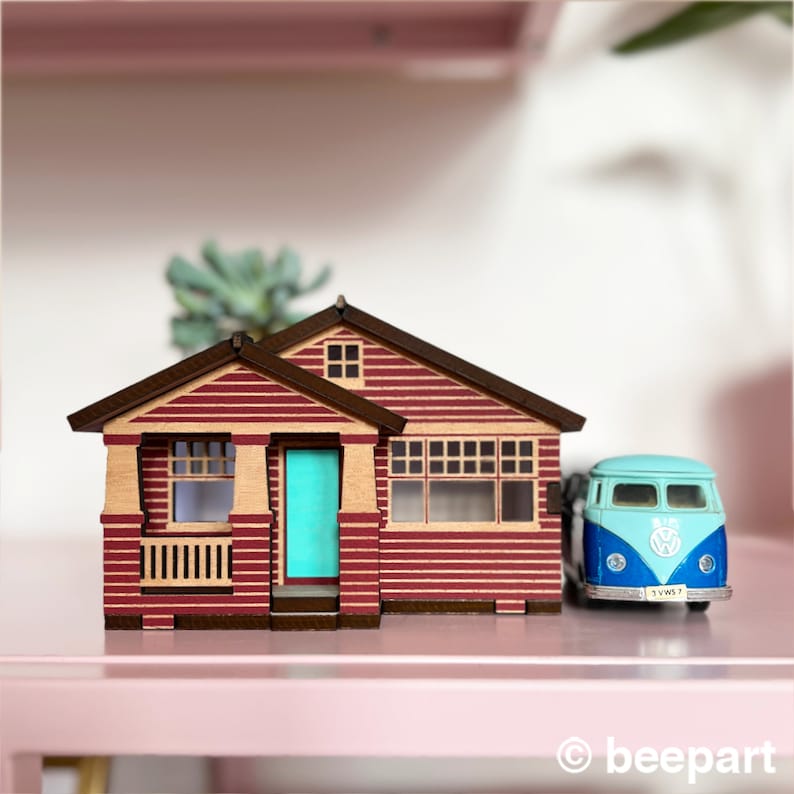 May include: A miniature wooden model of a red and brown house with a teal door and a blue and white Volkswagen van parked in front. The house has a porch with a railing and a window on the side. The van has a license plate that reads "1YWS7".