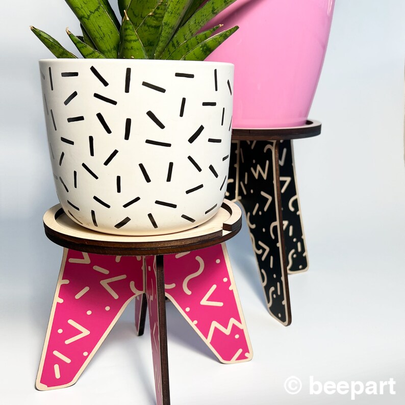 May include: A white planter with a black geometric pattern sits on a pink wooden plant stand with a geometric pattern. The stand has three legs and is made of wood. The planter is filled with a green plant.