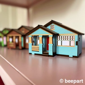 May include: A row of miniature wooden houses, with the closest one painted a light blue with a red door and brown trim. The houses are arranged on a pink surface.