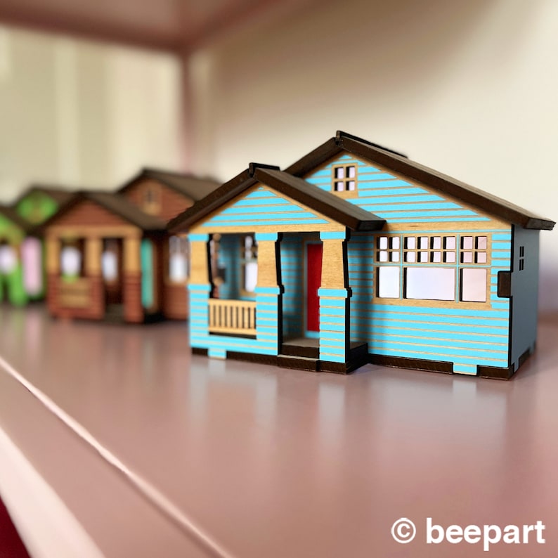 May include: A row of miniature wooden houses, with the closest one painted a light blue with a red door and brown trim. The houses are arranged on a pink surface.