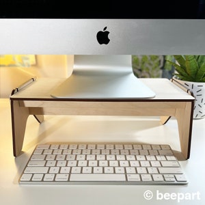 May include: A wooden computer monitor stand with a light wood finish and a dark brown edge. The stand is raising a silver computer monitor above a white keyboard.