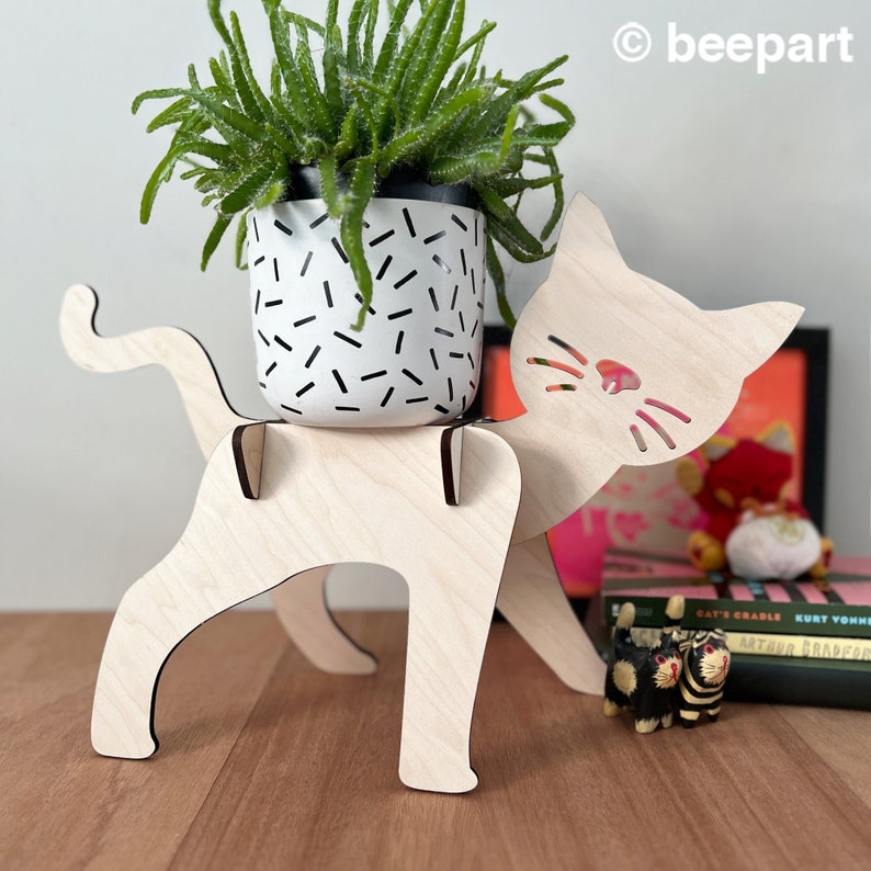 May include: Wooden cat planter stand with a white planter featuring a black confetti pattern, holding a green succulent plant. The cat stand has a cutout design for the cat's face and whiskers.