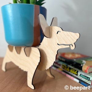 Könnte beinhalten: Ein hölzerner Hundepflanzenständer mit hellbrauner Oberfläche. Der Ständer ist in Form eines Hundes mit herausgestreckter Zunge geformt. Ein blauer und brauner Topf sitzt auf dem Ständer.