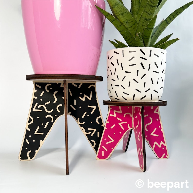 May include: Two plant stands with geometric patterns. The larger stand is black and white with a pink pot on top. The smaller stand is pink and white with a white pot with black geometric patterns on top.