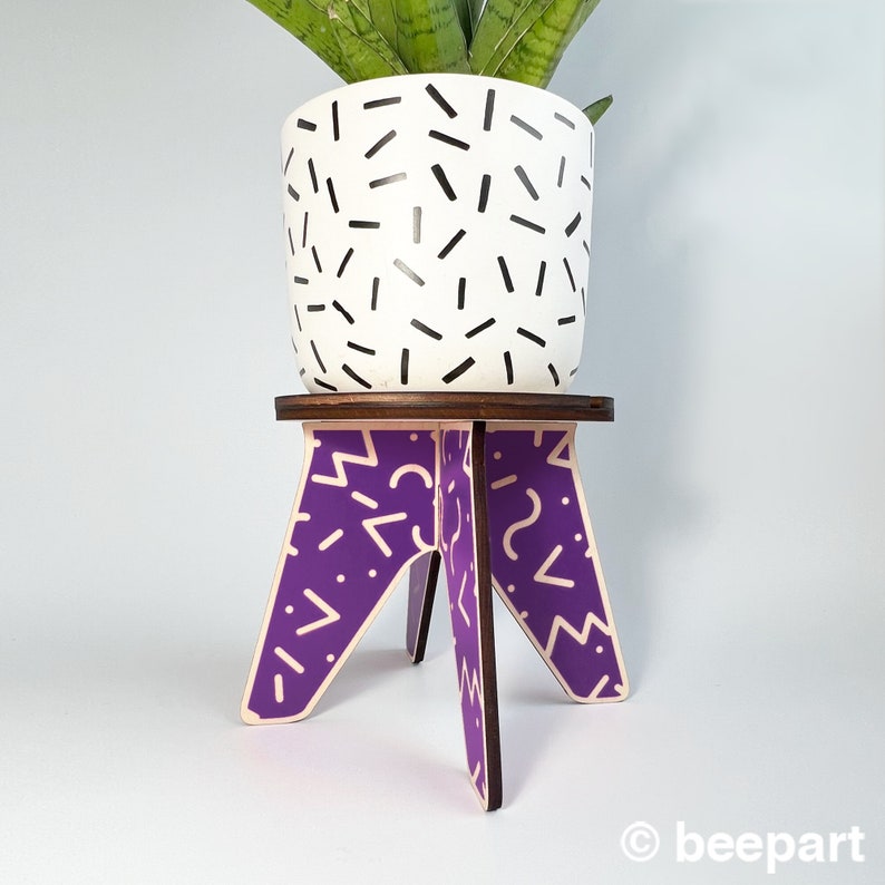 May include: A white planter with a black geometric pattern sits on a purple and white geometric patterned plant stand with three legs.