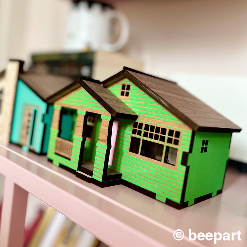 May include: A set of three miniature wooden houses with green siding and brown roofs. The houses are arranged in a row on a pink shelf.